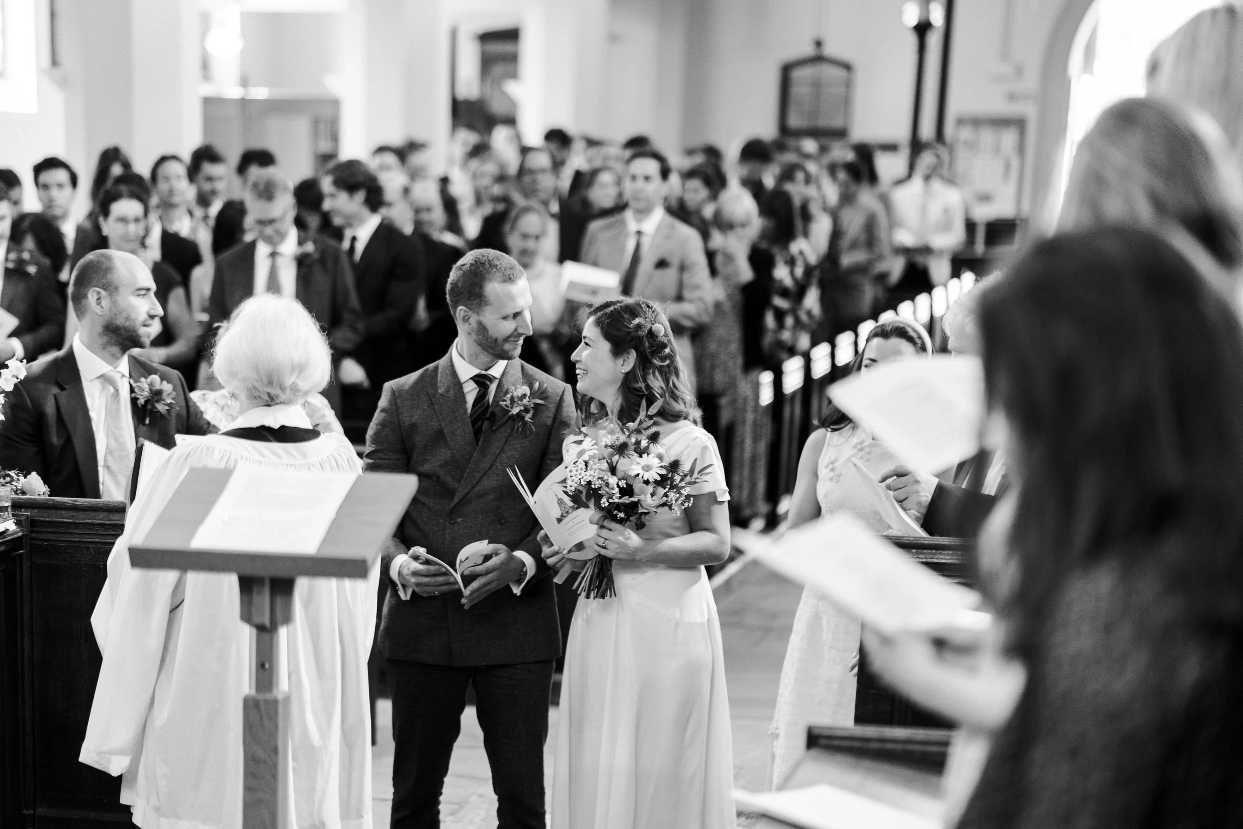 Wedding couple at the front of the altar looking and smiling at each other, while the choir who are just in focus at St Michael's Church at a Highgate, London Wedding
