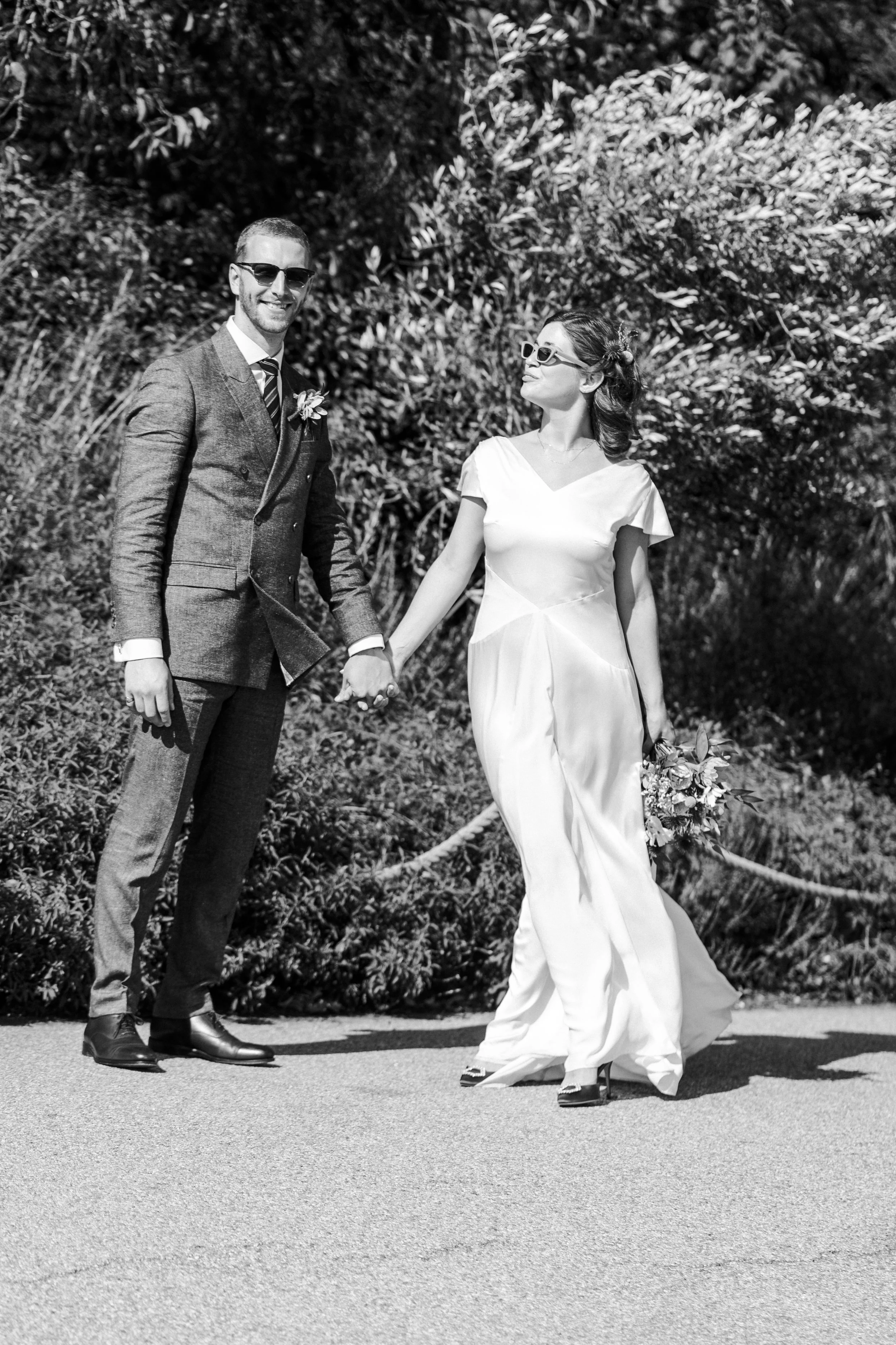 Wedding couple posing for portraits wearing sunglasses standing holding hands with groom looking and smiling at the camera and the bride looking at the groom  at a Highgate, London Wedding.