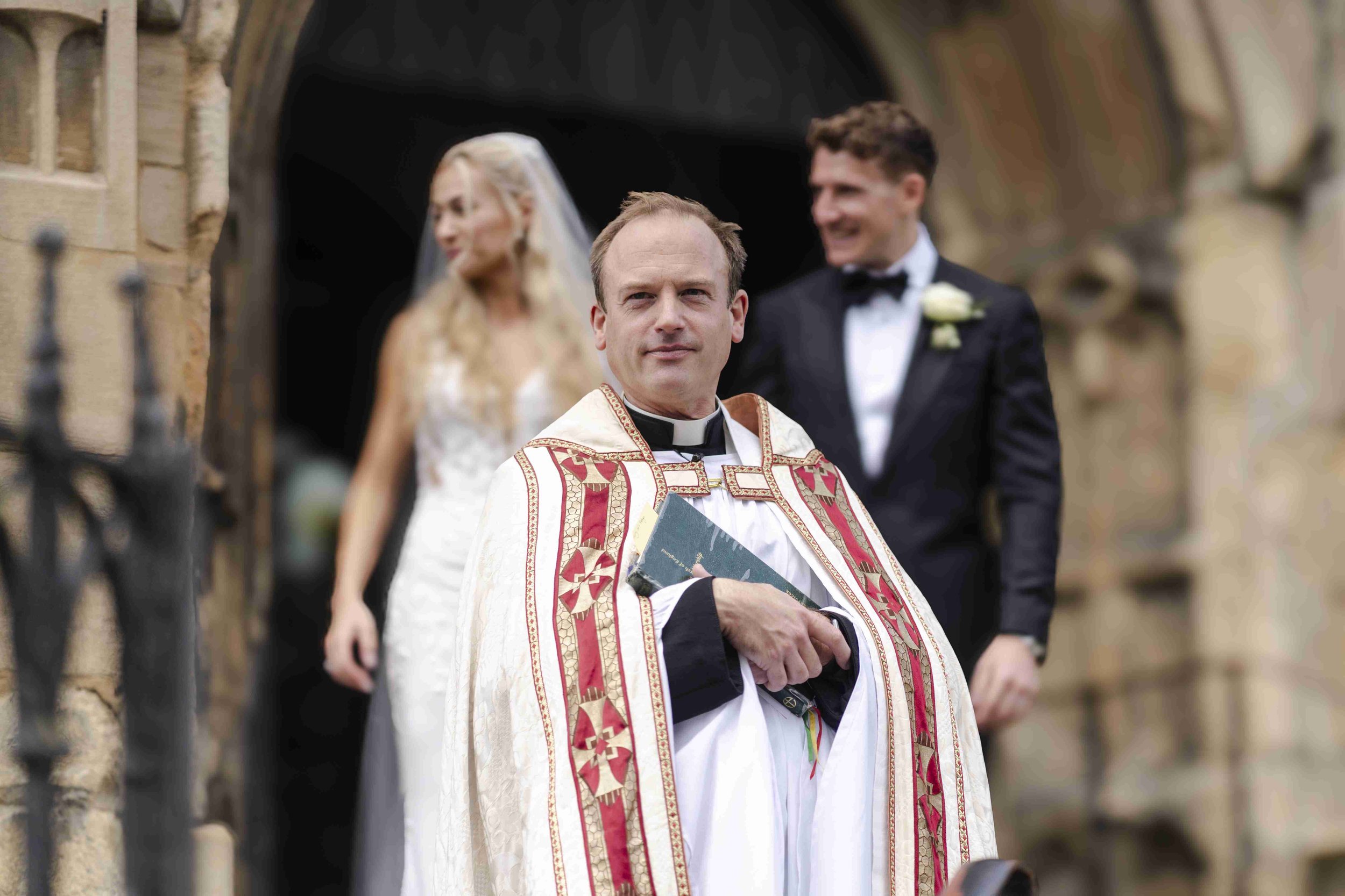 Vicar smiling at the camera standing outside the church with the couple in the background at the Halfmoon Farm Wedding in Rutland