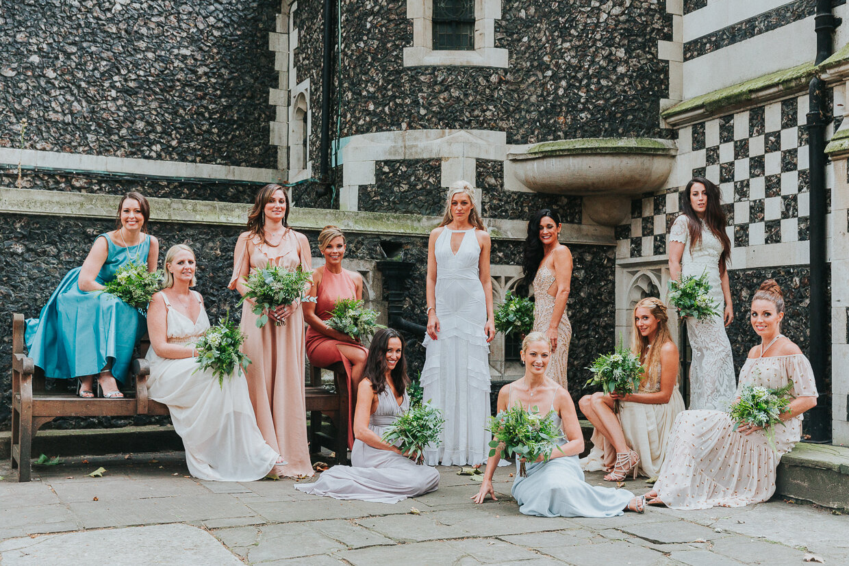 Group shot of a London Weddings Bride and her bridemaids in multicolour dresses standing and sitting outside St Bartholomew the Great Church in City of London