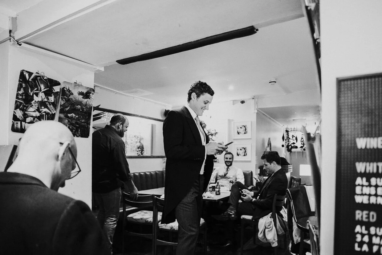 Groom standing next to a table using his phone, while guests sit around in the bar at a City of London Wedding.