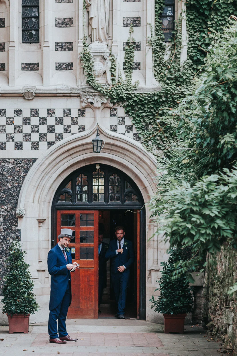 A Groomsmen stand outside the entrance to St Bartholomew the Great Church in Smithfield with a guest walking out the doorway at a City of London Wedding