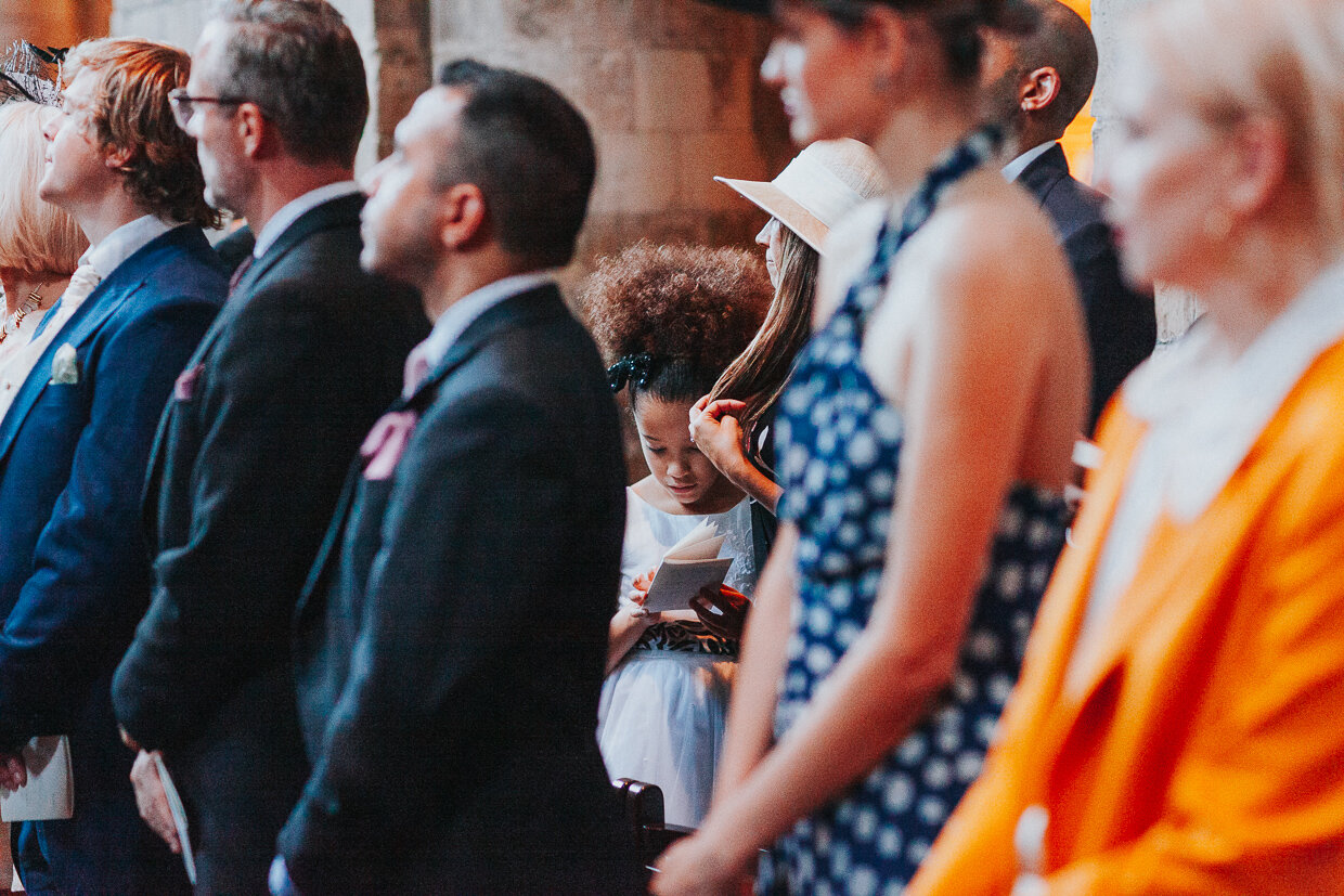 A view of a child guest, through guests in the forefront, looking at the Order of service at a City of London Wedding.