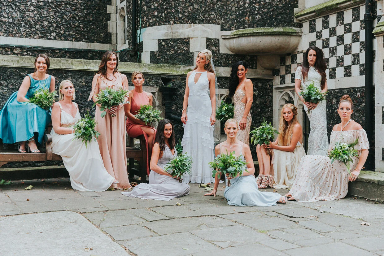 A group of women dressed in formal gowns holding bouquets of greenery and flowers, posing outdoors in front of a stone and brick background, with some sitting and some standing.