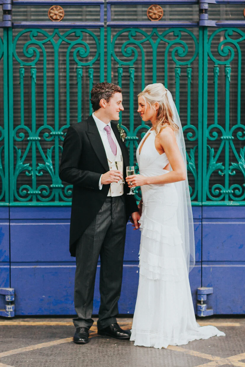 Bride & Groom standing next to metal gates in Smithfield Market looking at each other holding glasses of champagne at a City of London Wedding.