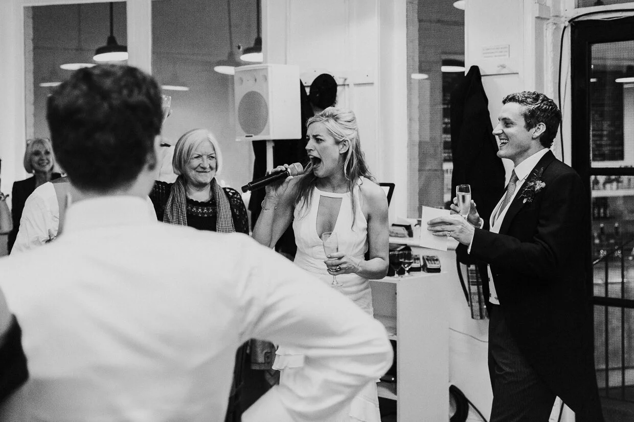 Bride at the reception, shouting into the microphone with Groom smiling standing next to her at St John Restaurant, Smithfield for a City of London Wedding.