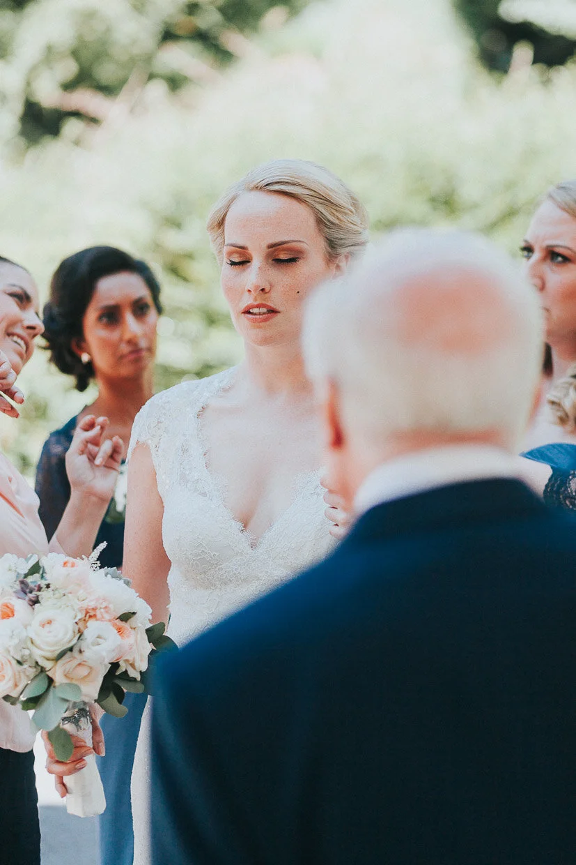 Bride outside the church with her eyes closed while a bridesmaid does final touch up on the Ballynatray Estate for a Cork Wedding