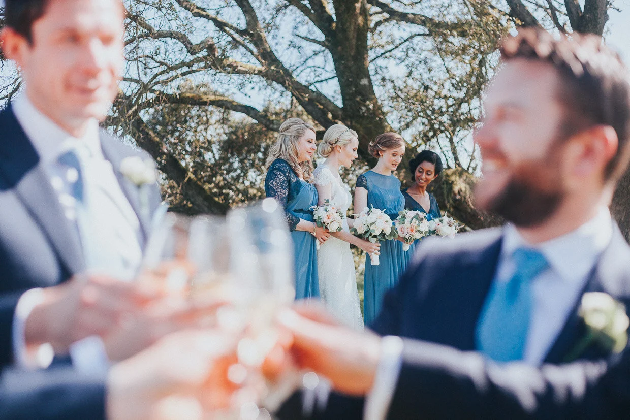 View of the bridal party in the background through blurred shot of the groomsmen toasting on the Ballynatray Estate for a Cork Wedding
