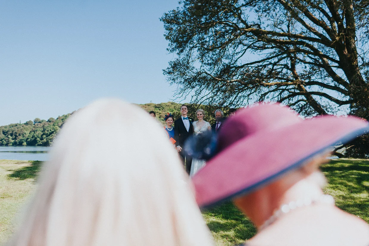 Bridal Party gathering for a group shot under a tree in the background with the shot taken through guests on the Ballynatray Estate for a Cork Wedding
