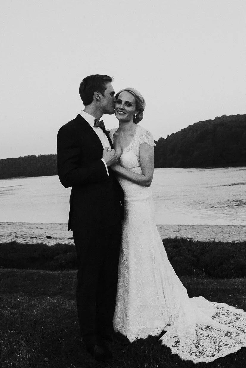 Groom kissing the Bride of the side of her head while posing next to the Lake on the grounds on the Ballynatray Estate for a Cork Wedding