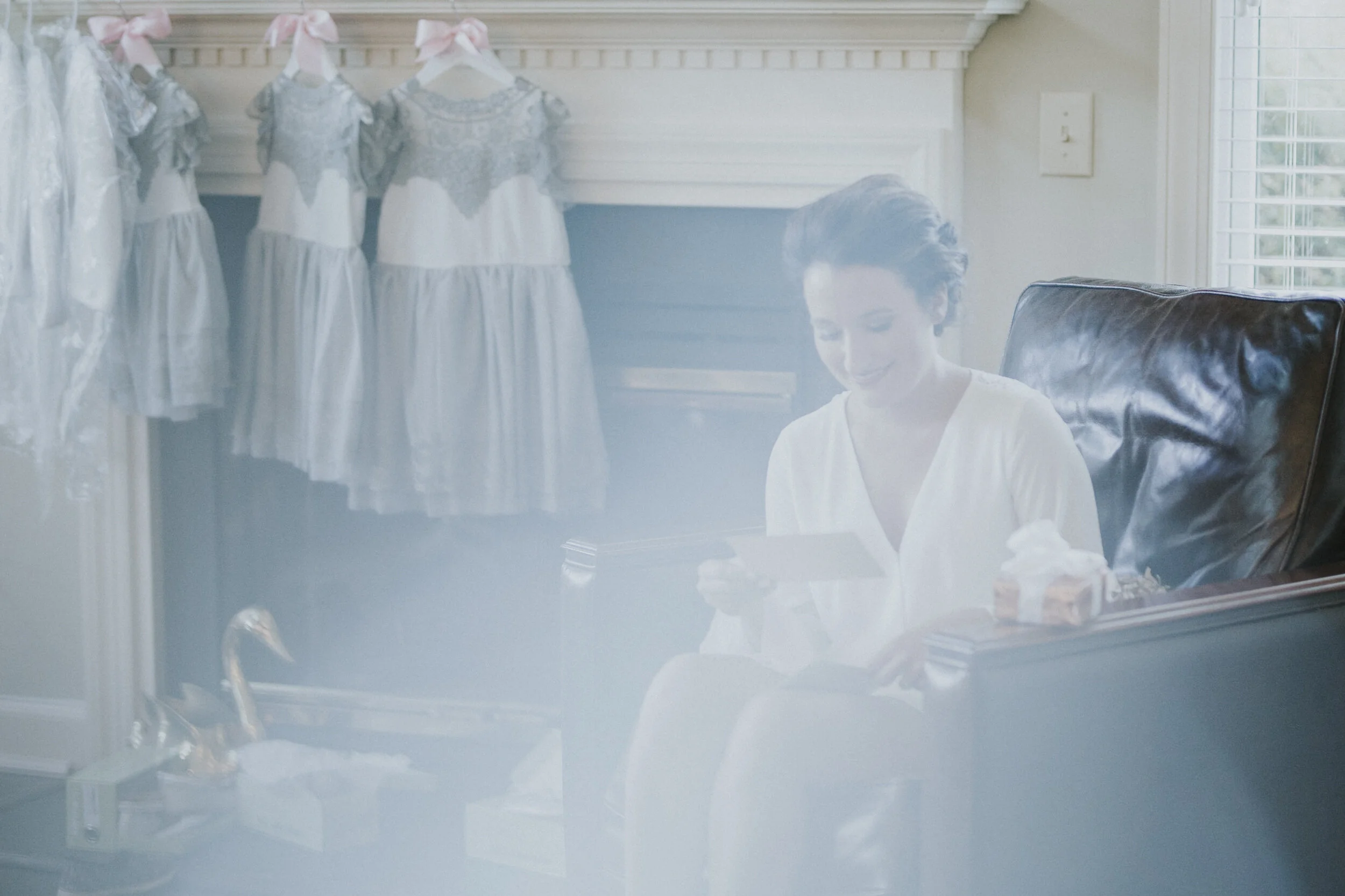 A International Bride sitting in an armchair reading a letter with bridesmaids dressing hanging behind her on the mantlepiece at a North Carolina Wedding.