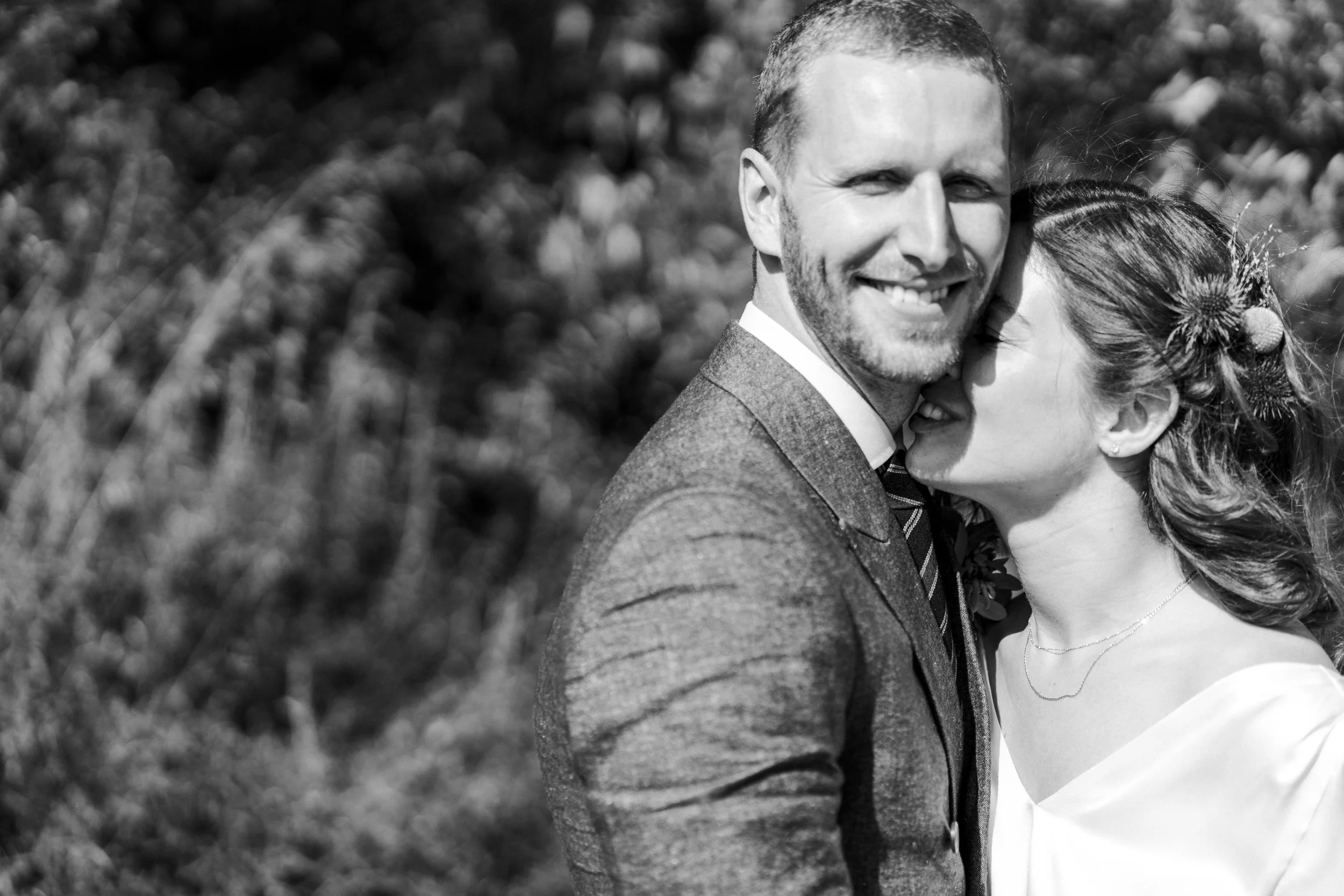 A smiling groom in a suit embraces his bride, who whispers in his ear outdoors—classic London Highgate Wedding Photography.