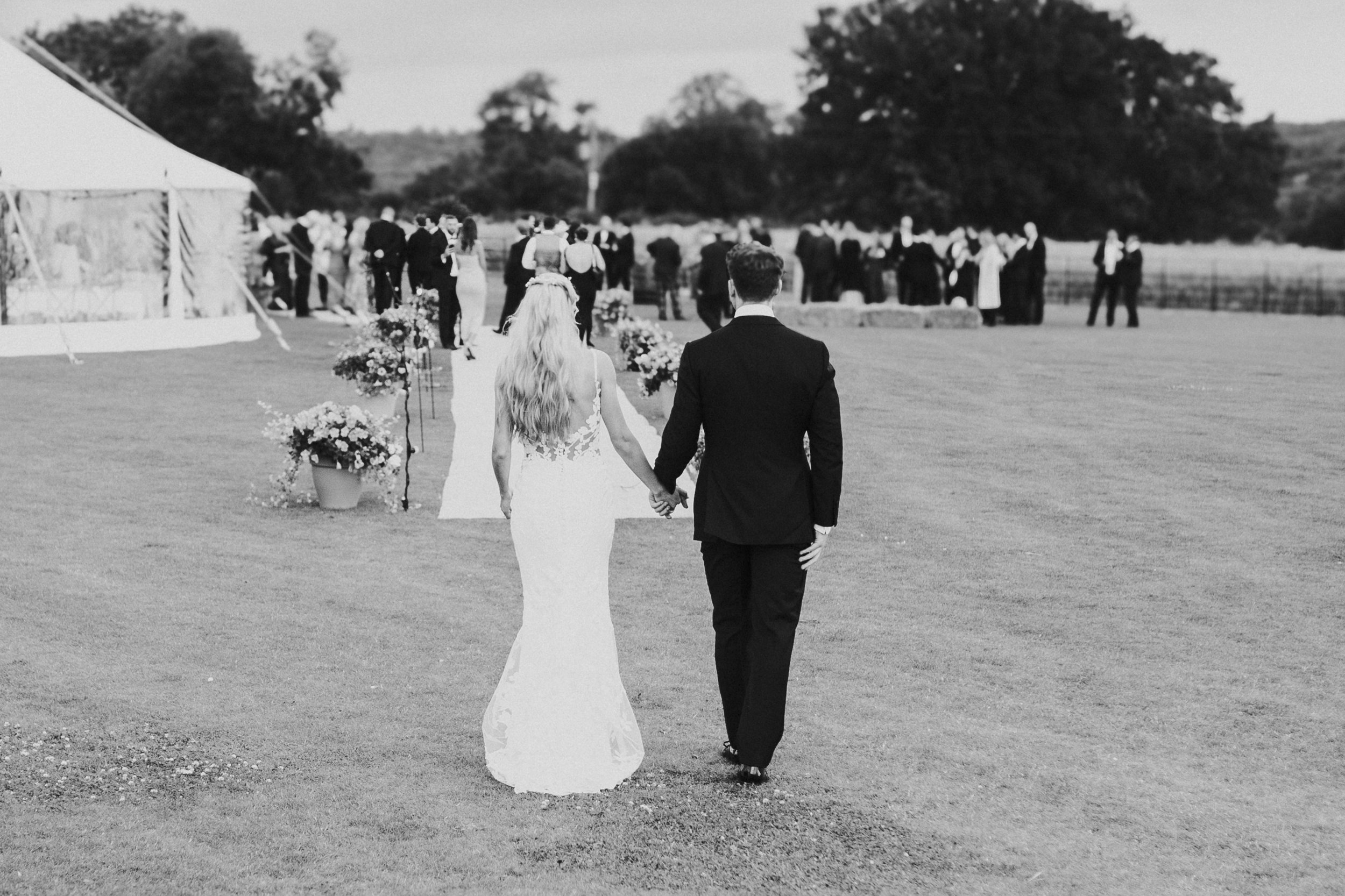 A bride and groom walking hand in hand on a wedding aisle outdoors, with the bride in a white dress and the groom in a black suit, leading to a group of people gathered near a tent in the background.