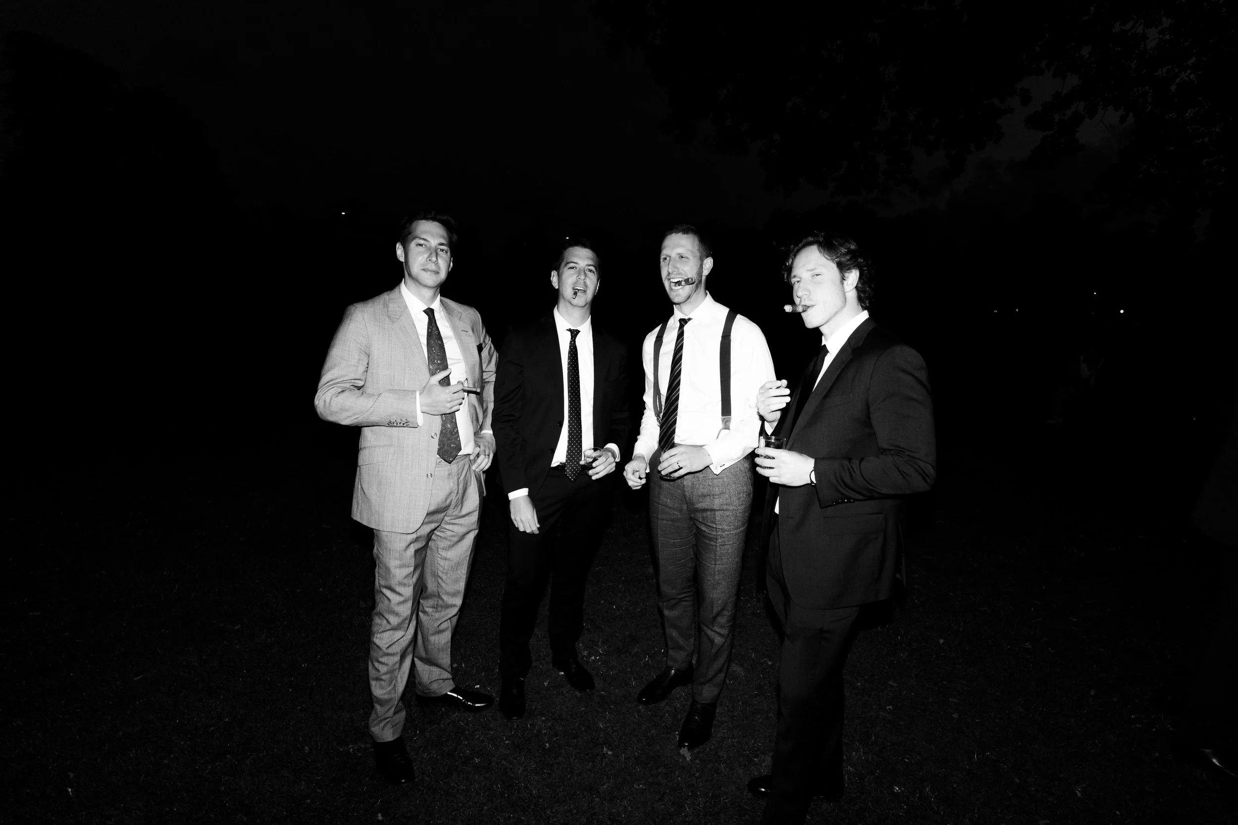 Groom with guests holding drinks and cigars with harsh flash lighting them at a Highgate, London Wedding.