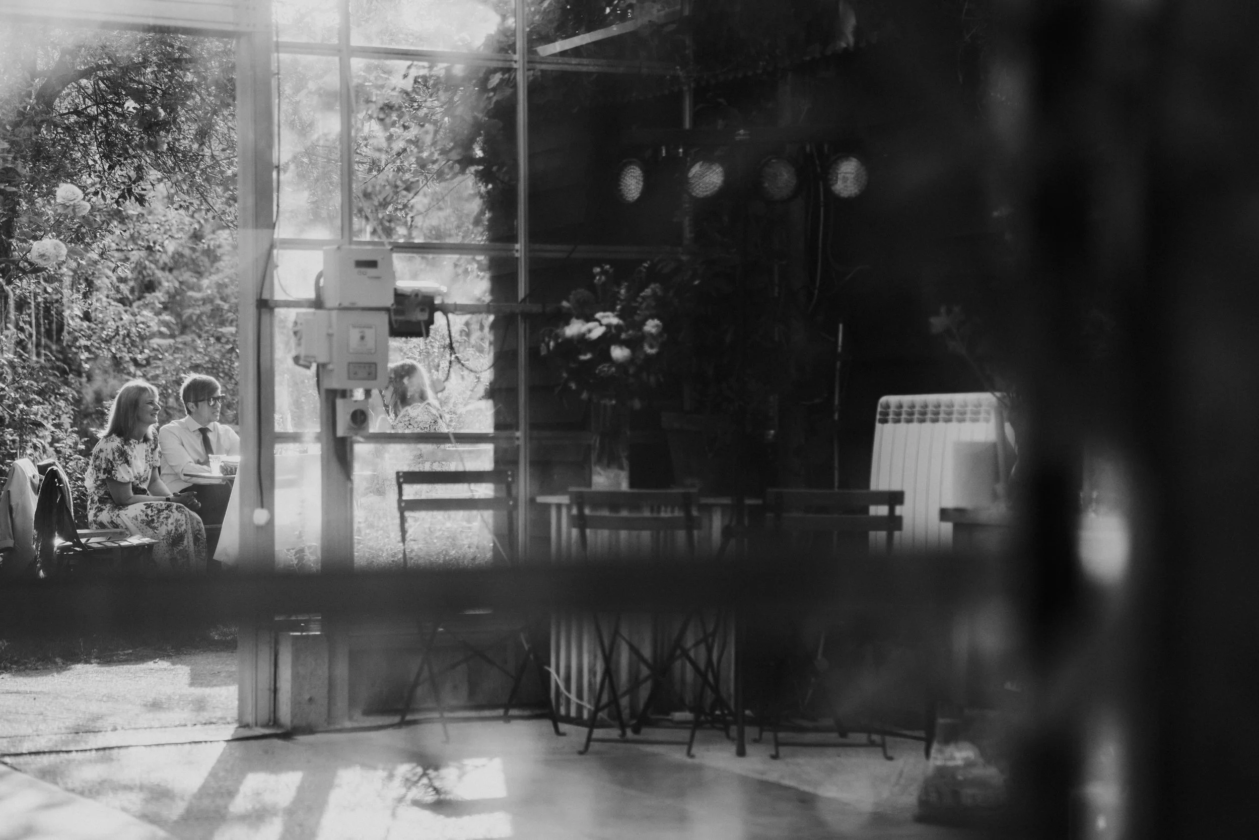 View through Greenhouse glass panels of wedding guests sitting outside wedding reception room at a Worton Kitchen Garden, Oxfordshire Wedding.