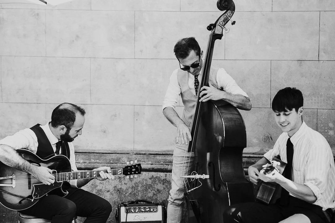 A 3 piece band with 2 guitarist and a cello player, smiling and playing at a Hampton Court House Wedding.