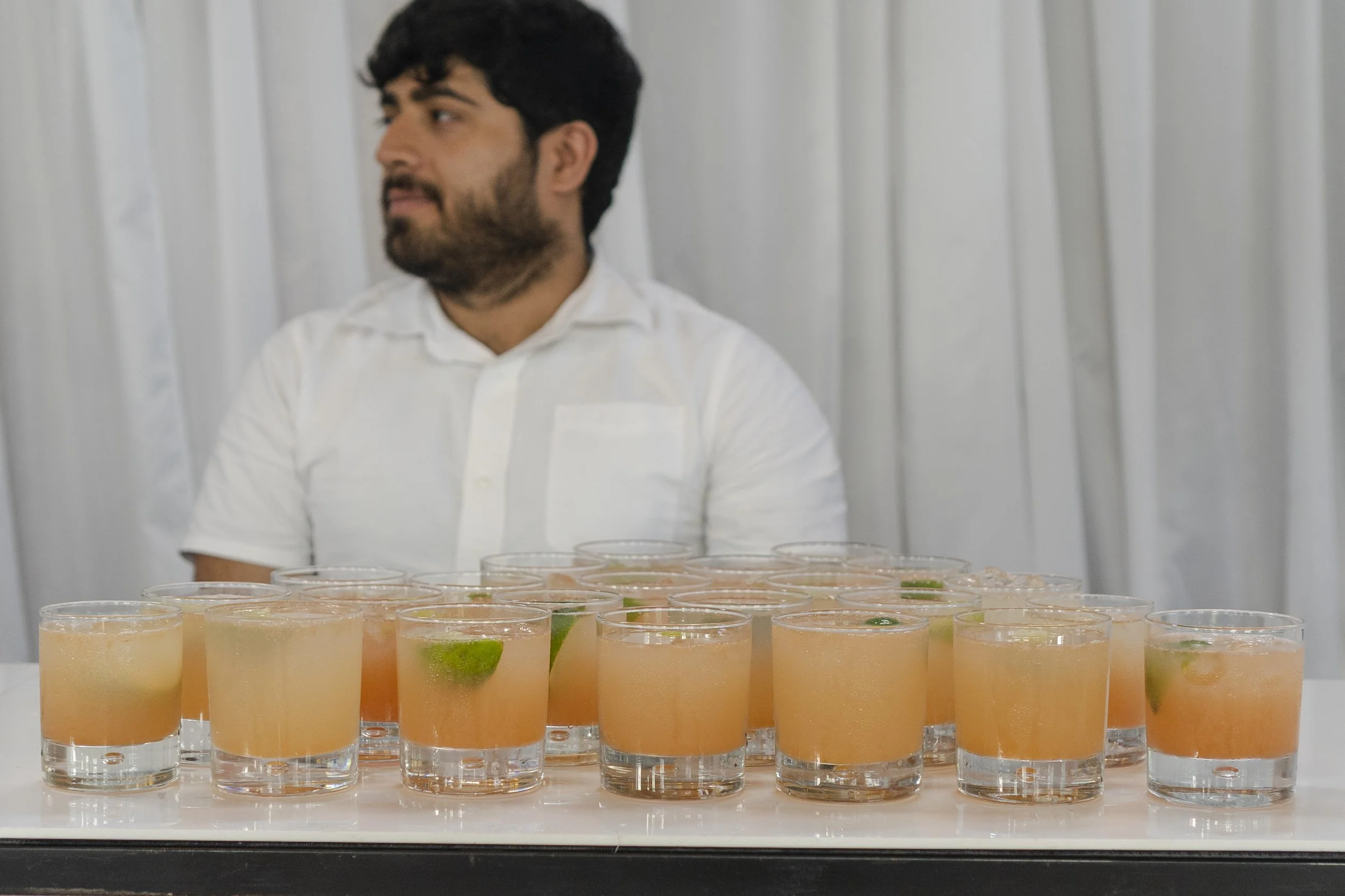 Waiter behind rows of glasses with cocktails Wedding couple posing in Highgate School in front of large artwork slightly back to back at a Highgate, London Wedding.