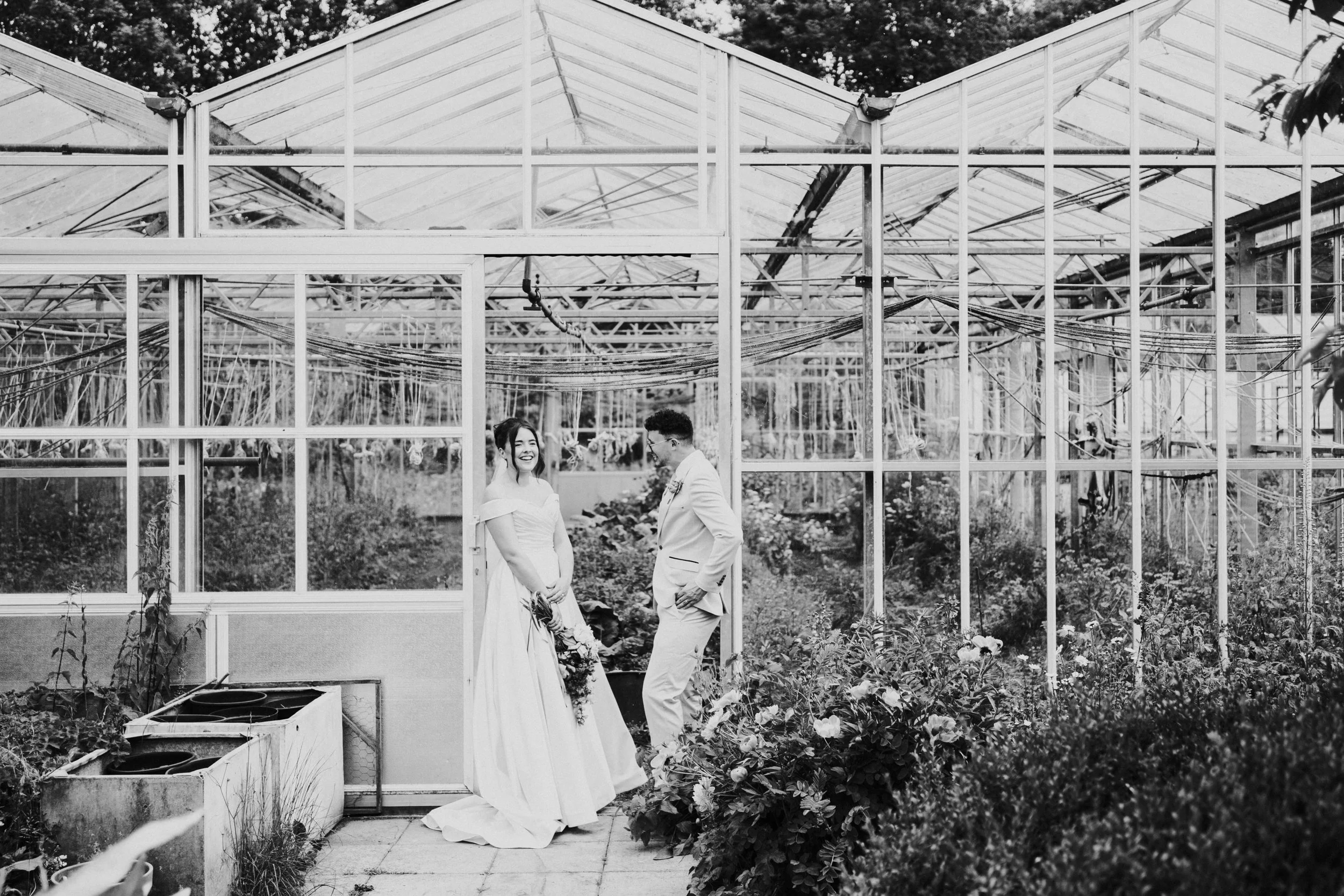 Wedding couple leaning against a doorframe to a greenhouse with the bride looking and smiling at the camera while the groom looks at the bride at a Worton Kitchen Garden, Oxfordshire Wedding.
