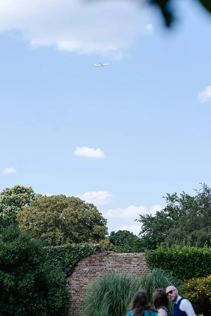 A view of the sky above a wall at Hampton Court House with a plane in the distance with a few guests in the bottom of the image at a London Wedding.
