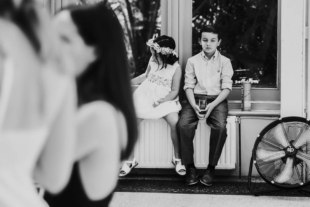 Two young guests with one wearing a flower crown sitting on the windowsill with a tube of Pringles next to them at a Hampton Court Wedding.