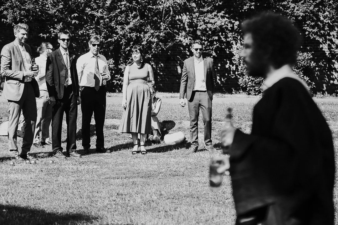 Guests mingling around on Hampton Court House's Lawn in the background while a blurred guest in the foreground walks past the camera at a Hampton Court Wedding.