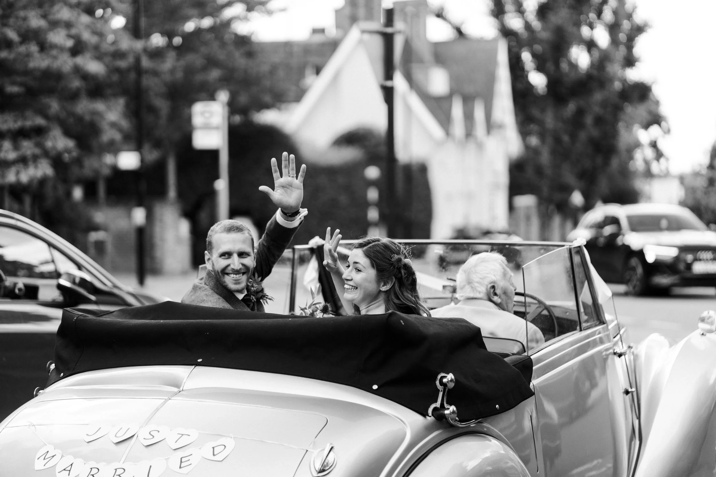 Wedding couple at the back of wedding car with the top down waving backwards to guest while being driven away at a Highgate, London Wedding