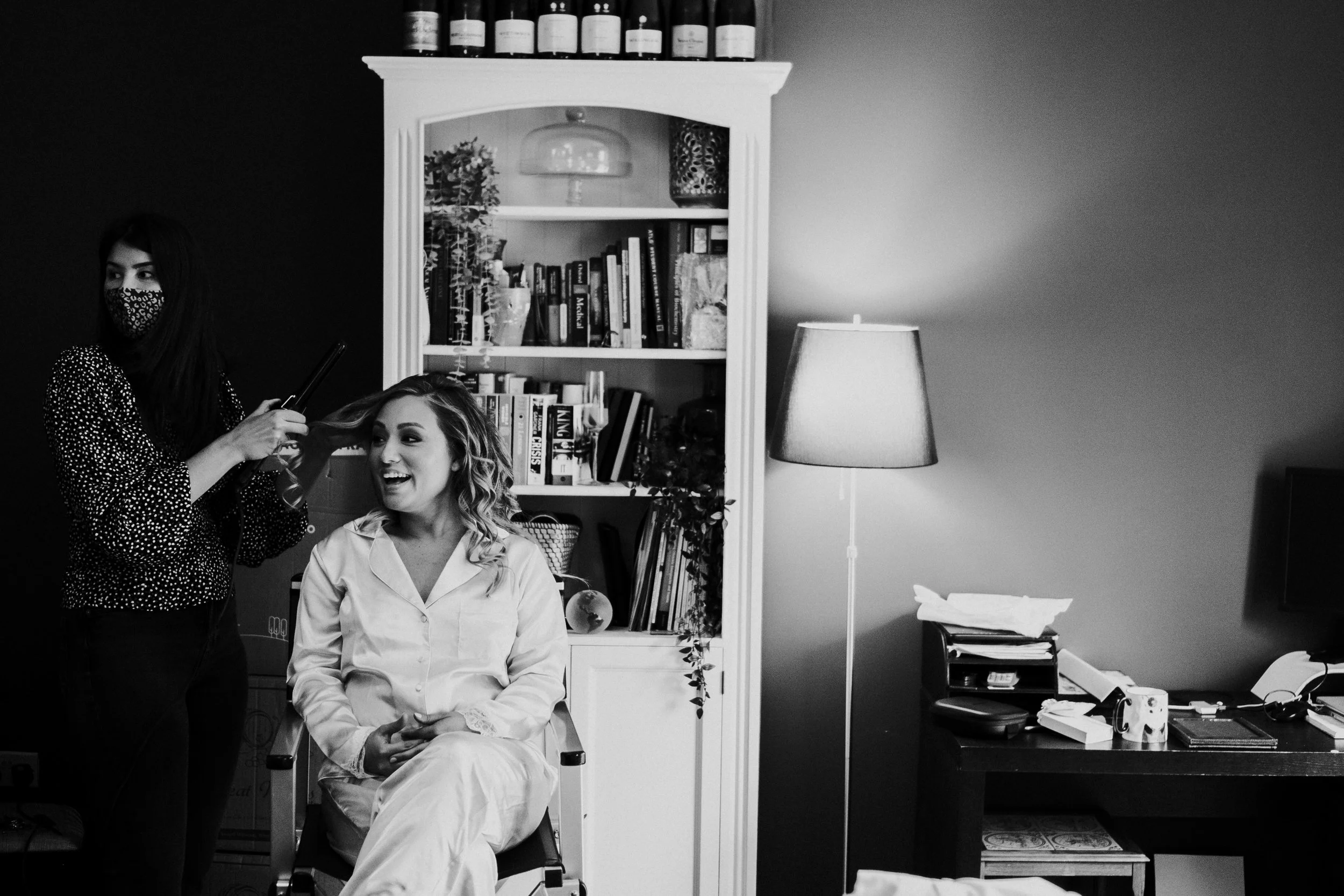 Bride smiling and laughing with someone out of shot, while the hairdressers who also is looking at someone out of shot is holding the brides hair with curly togs in her other hand at a Pembroke College Oxford University Wedding