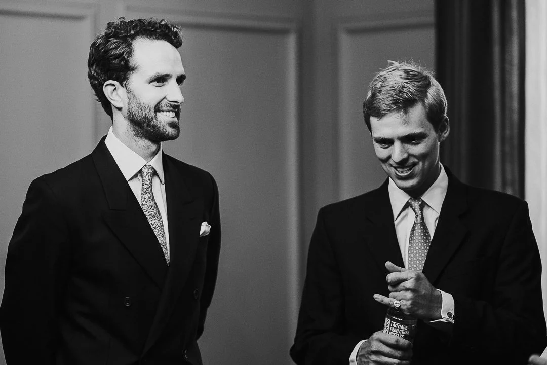 Groom smiling while a guest who is holding a bottle is looking down and smiling in the ceremony room at the Old Marylebone Town Hall Wedding.