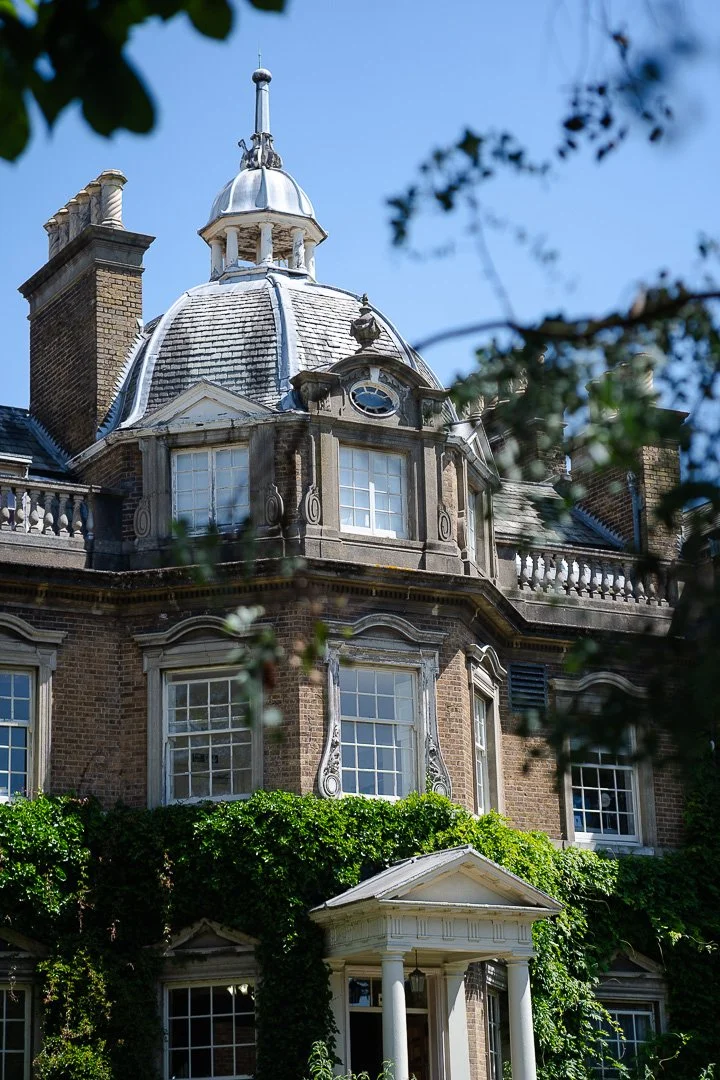 A side view of the entrance to Hampton Court House with foilage in the foreground of the image on a clear sunny day
