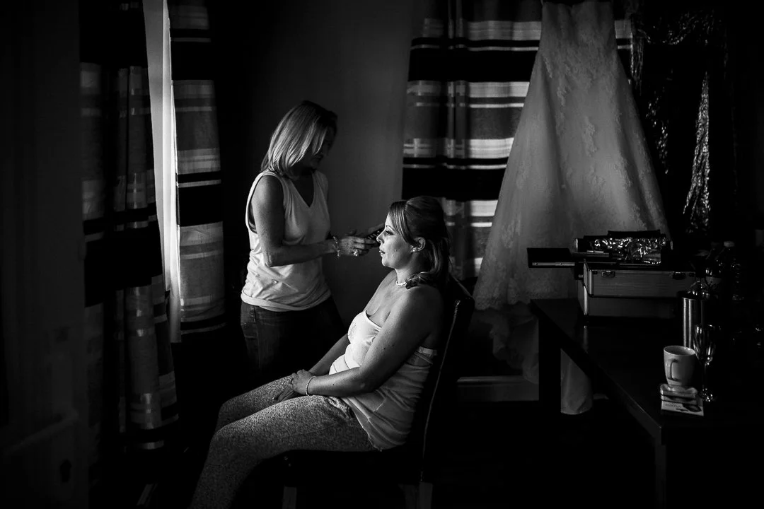 A woman getting her makeup done by a makeup artist in a dimly lit room with patterned curtains and a wedding dress hanging on the wall.
