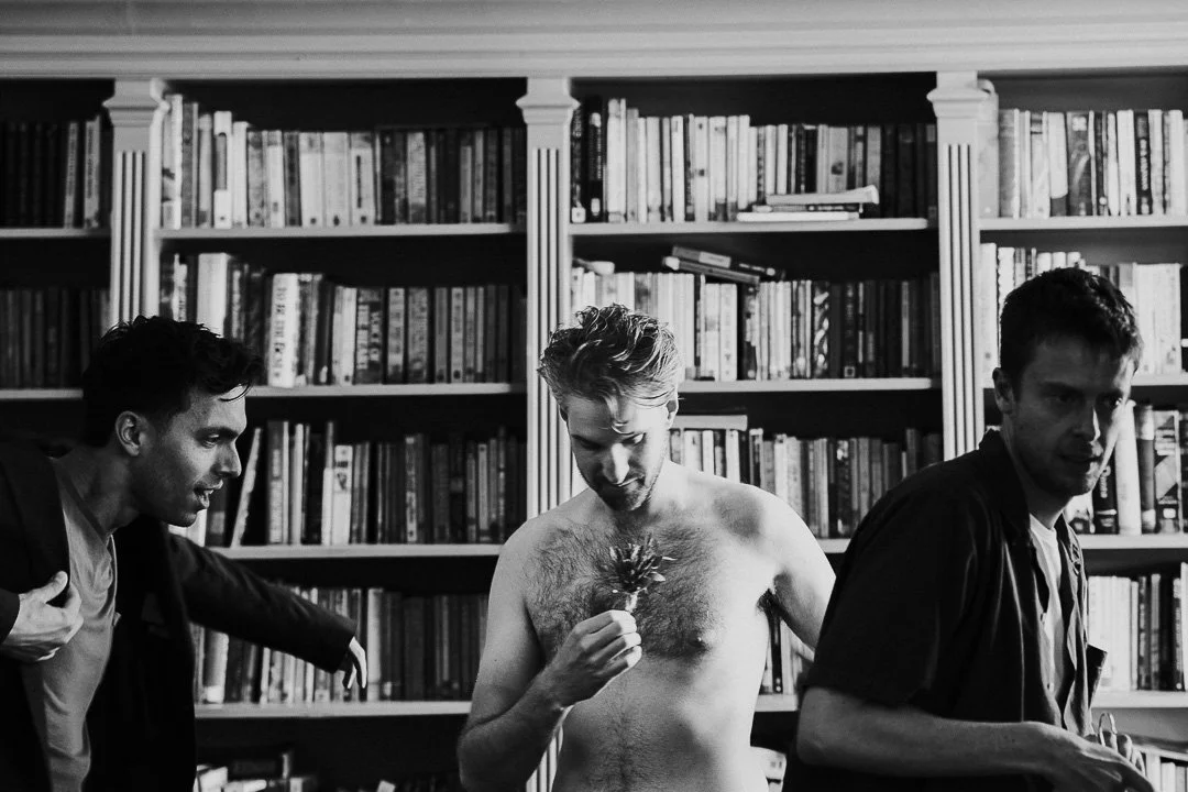 Groom with groomsmen in the library at Hampton Court House, bare chested holding a buttonhole towards his chest.