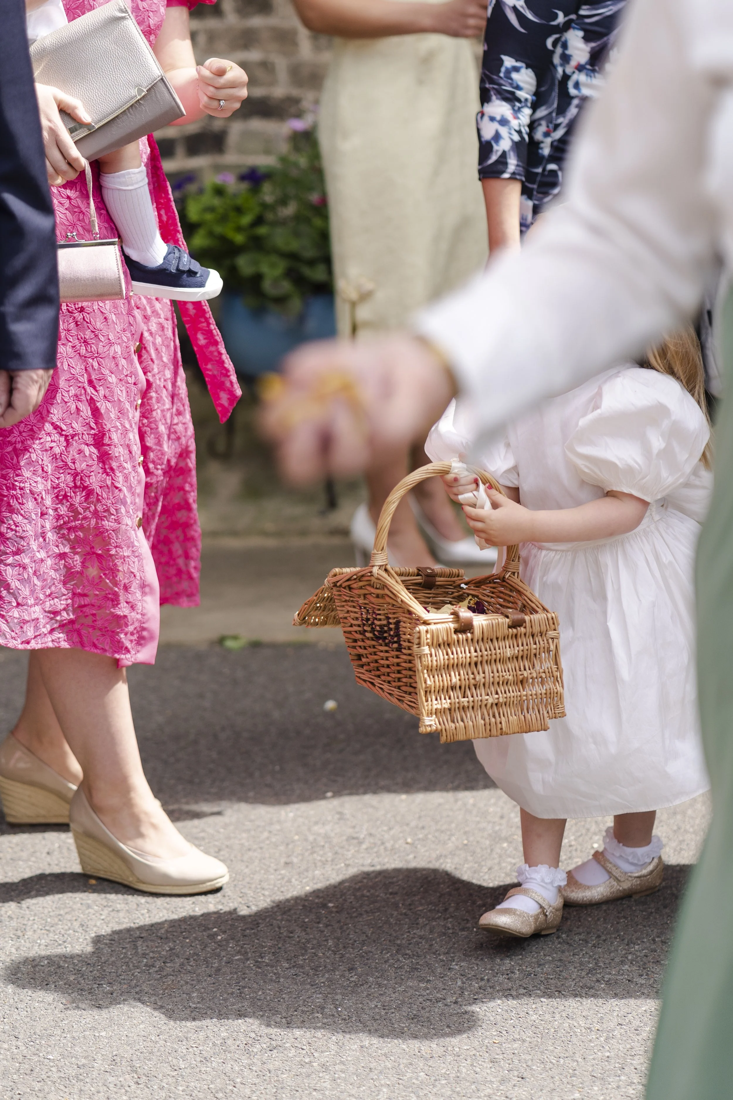 Flower girl with basket outside St Michael's Church with her head hidden behind a guest's arm at a Highgate, London Wedding