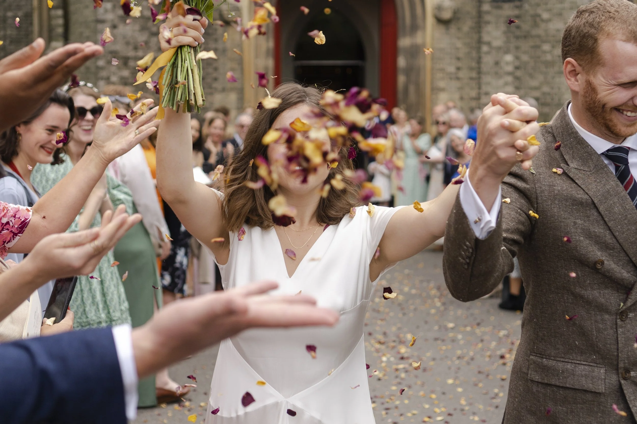Wedding couple walking down the confetti line with a guest throwing flower petals that covers the bride face at St Michael's Church at a Highgate, London Wedding