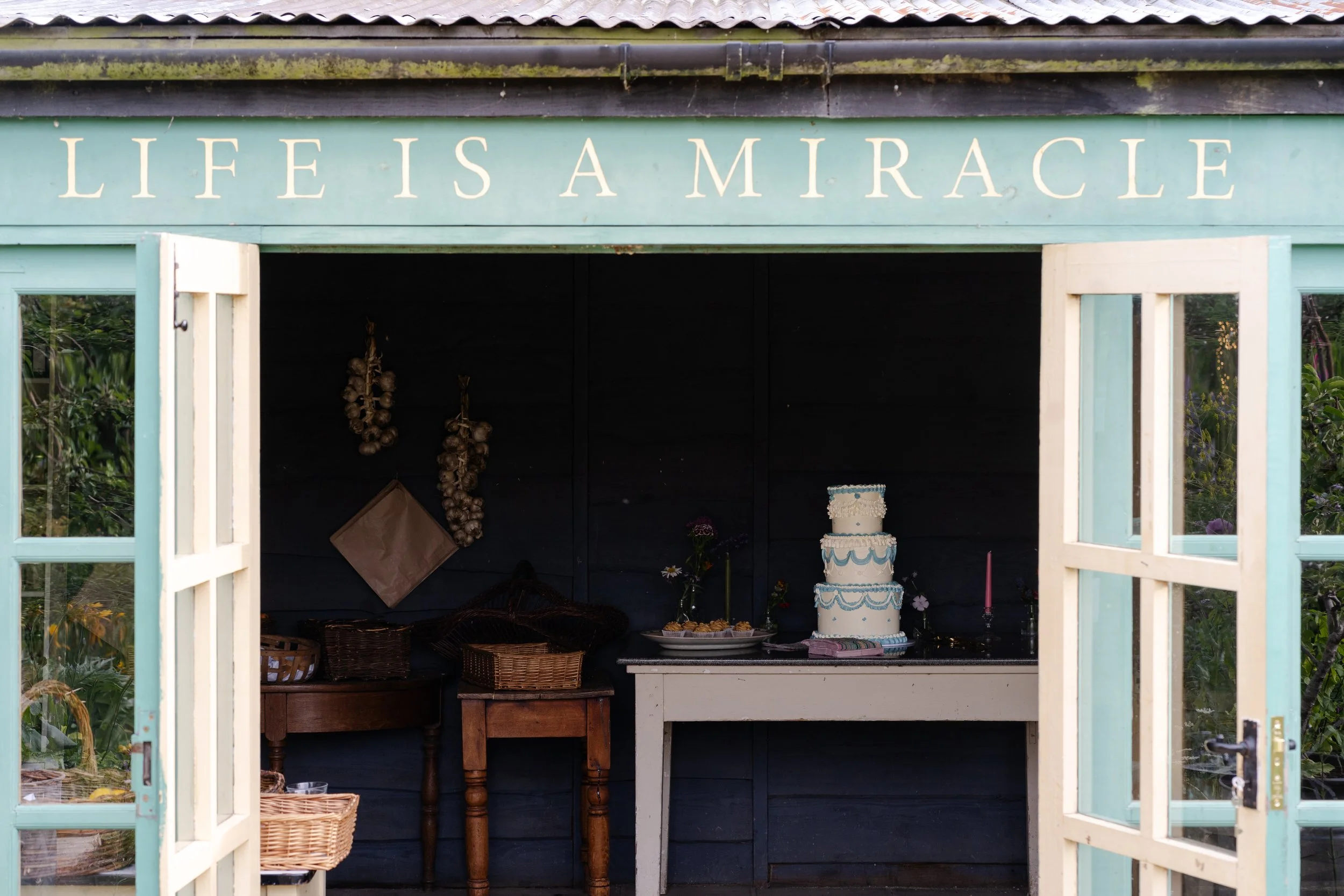 Wedding cake inside Hut with door opens under a sign saying Life is a Miracle at a Worton Kitchen Garden, Oxfordshire Wedding.