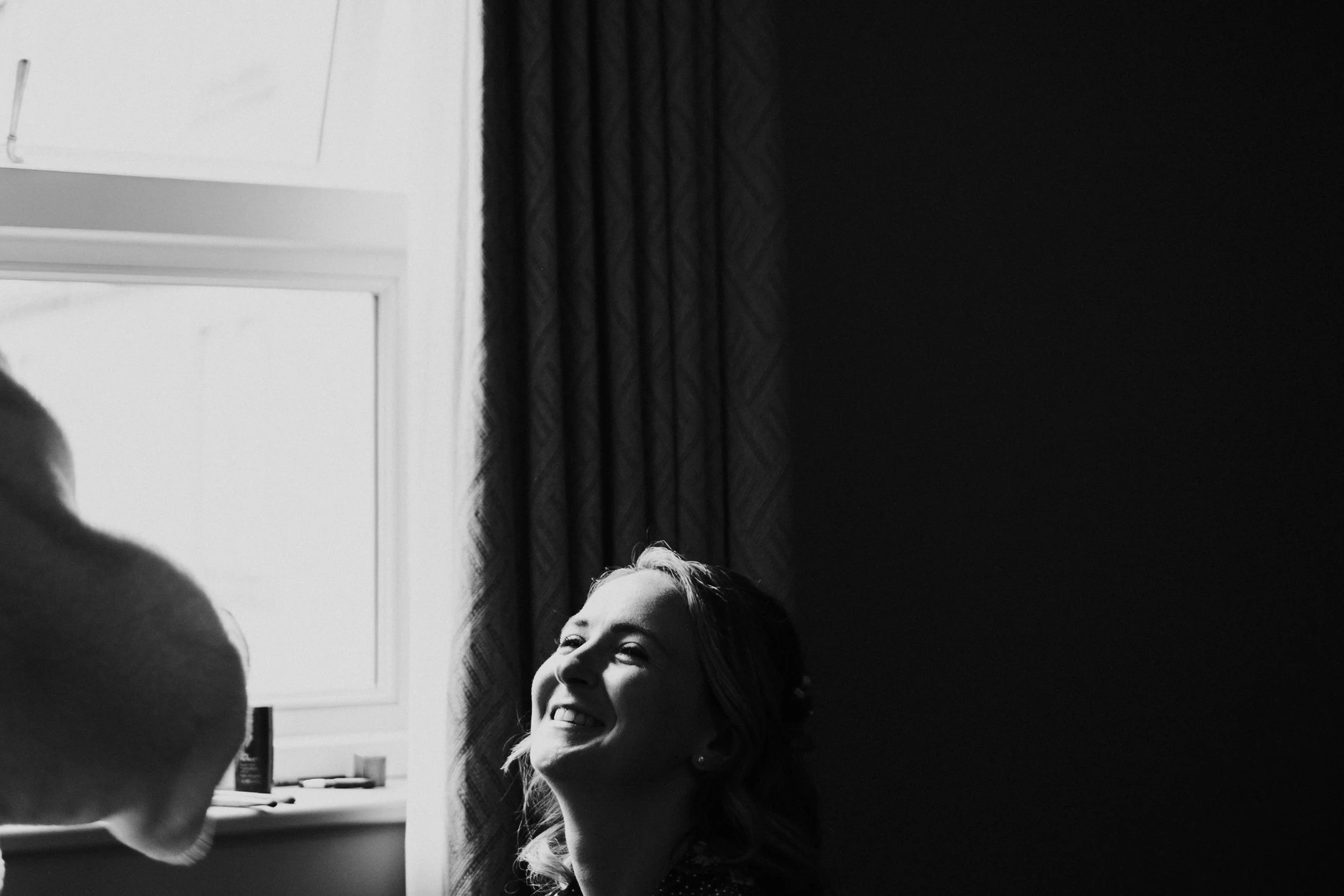 Bride looking up at her bridesmaid and smiling while sitting down with the window light shining on the right side of her face at a Oxford Town Hall Wedding