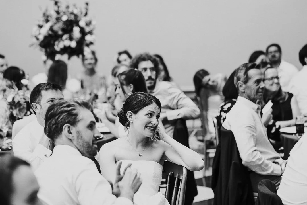 The bride sitting down at the dinner table watching and smiling at the speeches at a Hampton Court House Wedding
