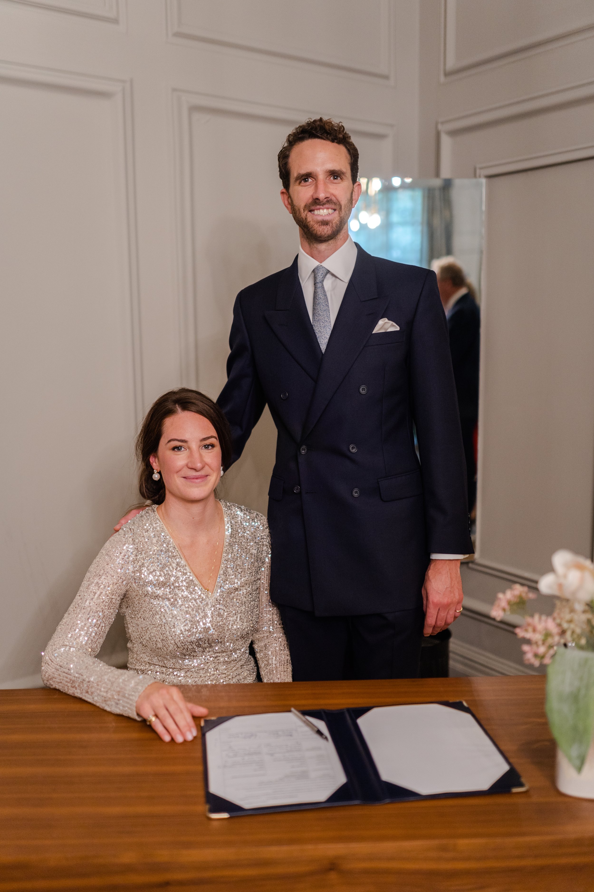 Wedding couple posing after signing the wedding registry, with the bride sitting down and the groom standing, both smiling at the camera in the ceremony room at the Old Marylebone Town Hall Wedding.