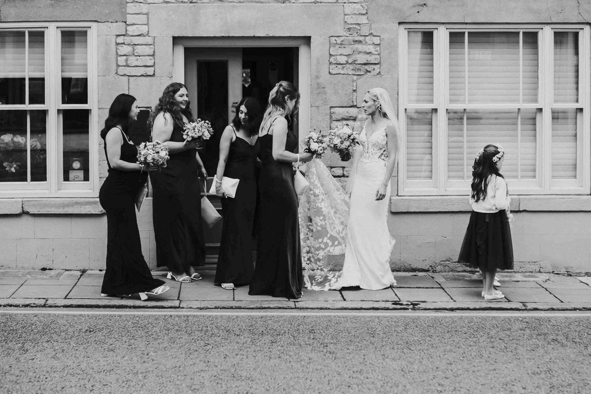 Bride stopping to talk to her bridal party while the flower girl walks on down the street to the church at the Halfmoon Farm Wedding in Rutland