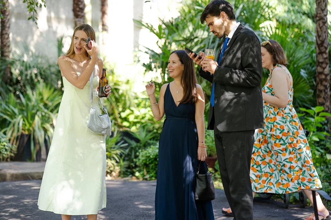 Three guests standing around with one on his phones while holding a drink while the other two are smiling and laughing slightly at a Hampton Court House Wedding
