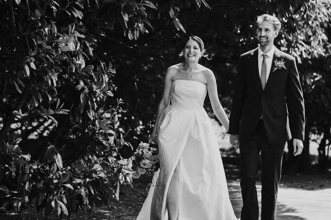 Bride & Groom walking through the gardens holding hands and smiling at a Hampton Court House Wedding.