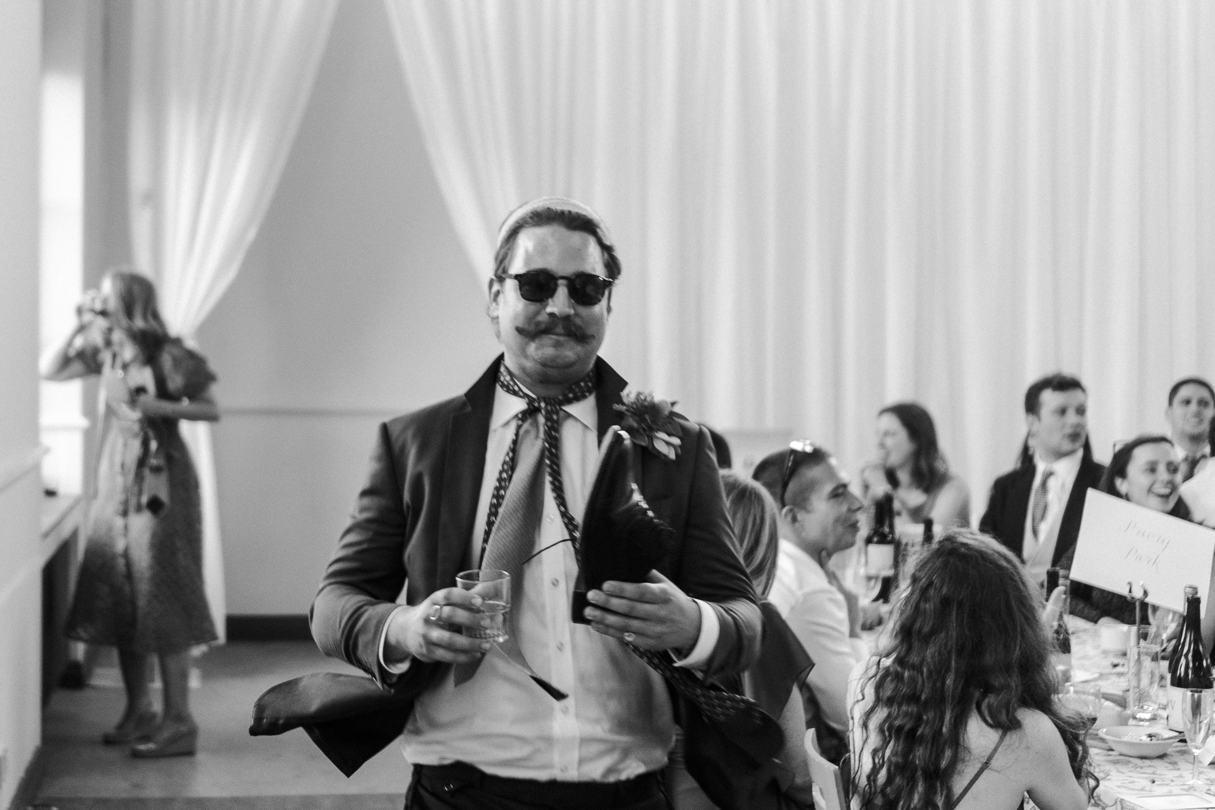 Wedding guest with a moustache wearing sunglasses and holding a glass and a shoe at a Highgate, London Wedding.