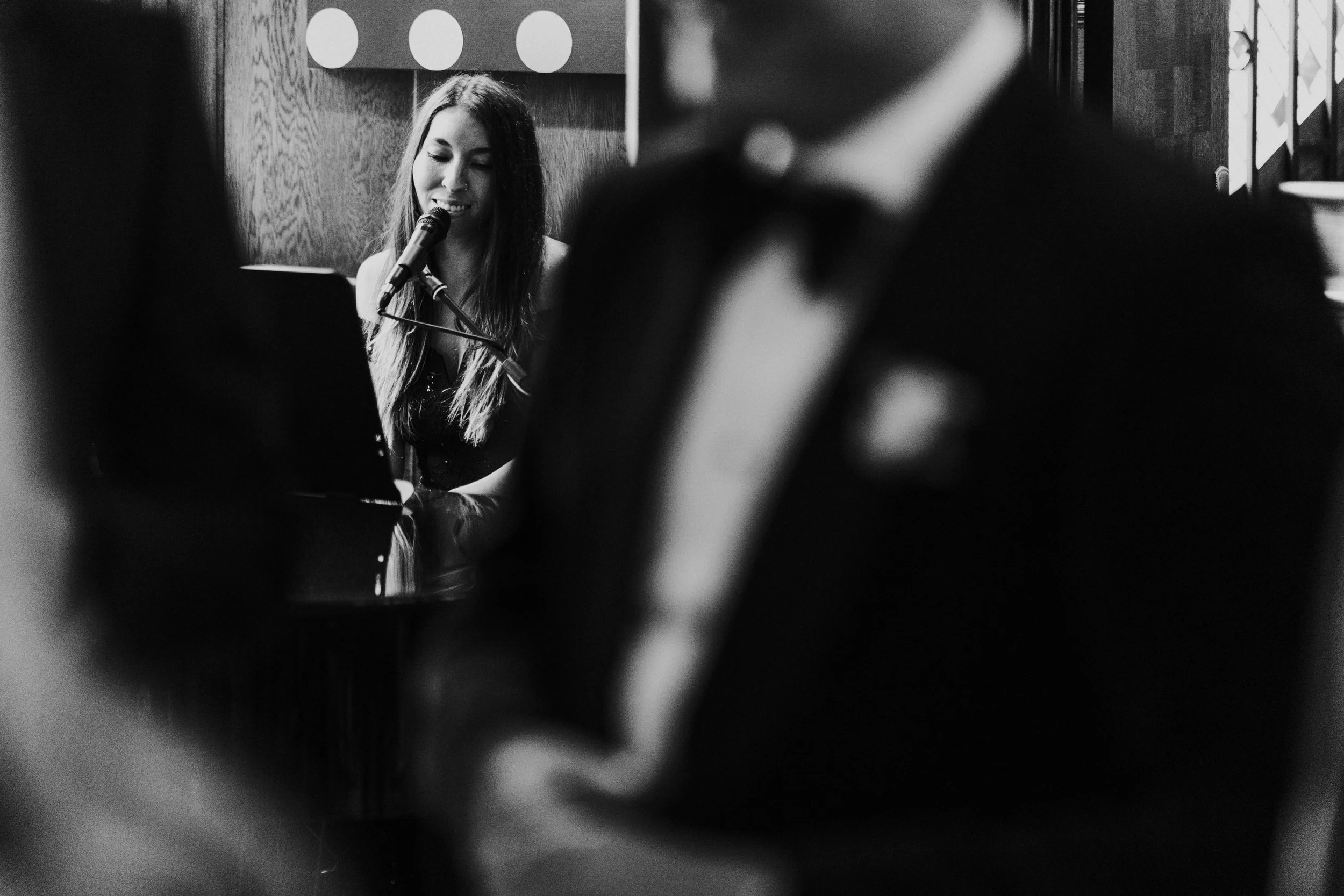 Wedding singer at the piano playing it, viewed through guests mingling at The Ivy Restaurant at a Old Marylebone Town Hall Wedding.