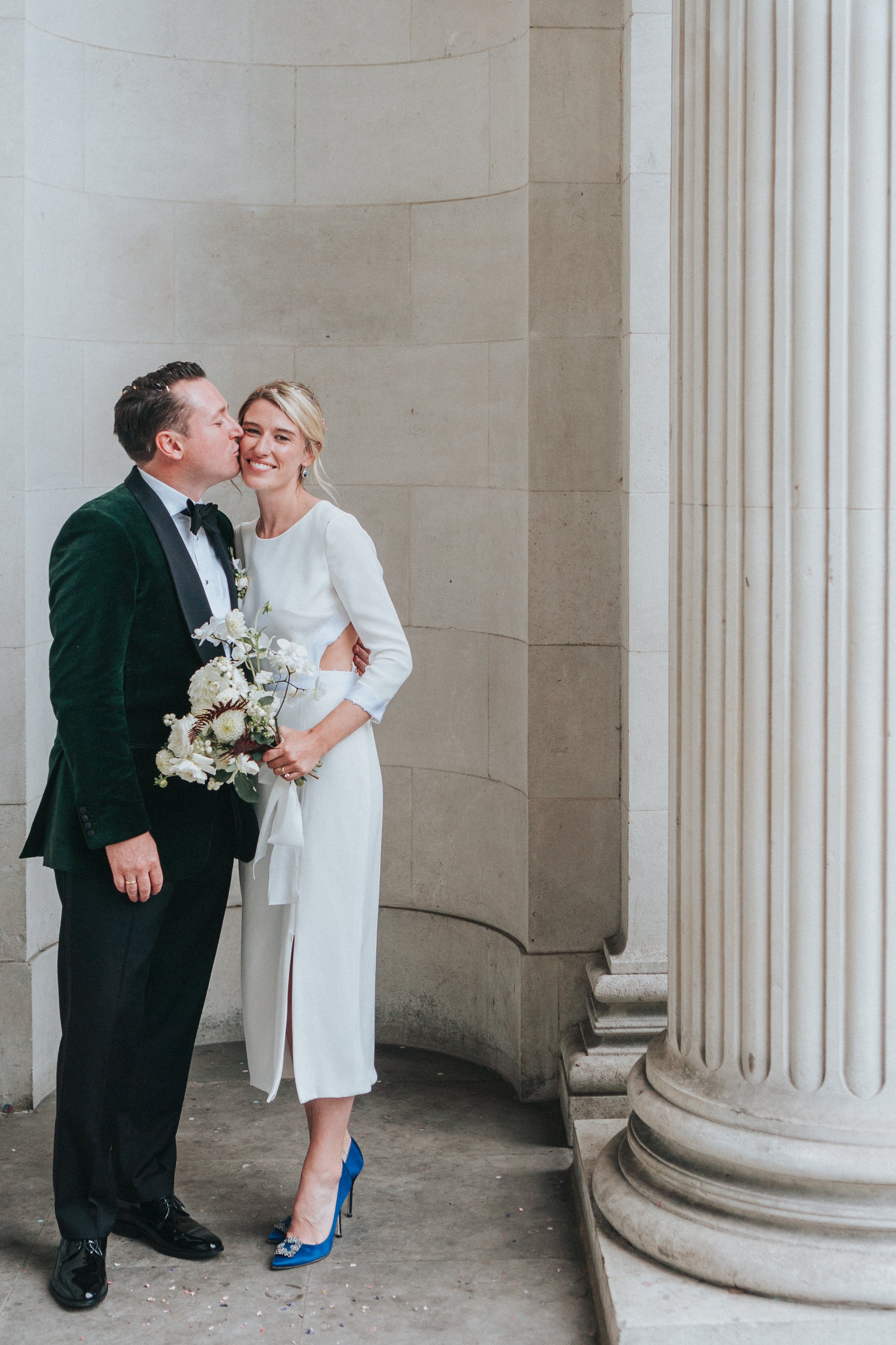 Couple posing for portraits next to the pillars with Groom kissing the Bride on the side of the head at a Old Marylebone Town Hall Wedding