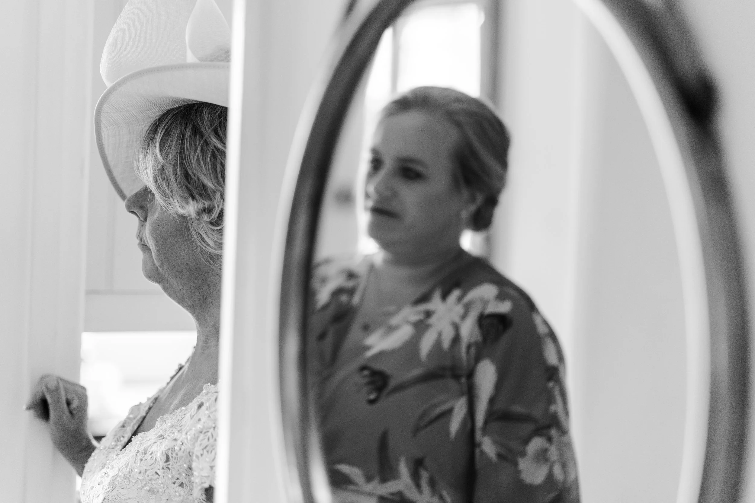 An image of the Bride's sister in law reflection in the mirror while the bride's mum is standing the other side of the wall just coming into shot at Earth Trust, Oxfordshire Wedding.