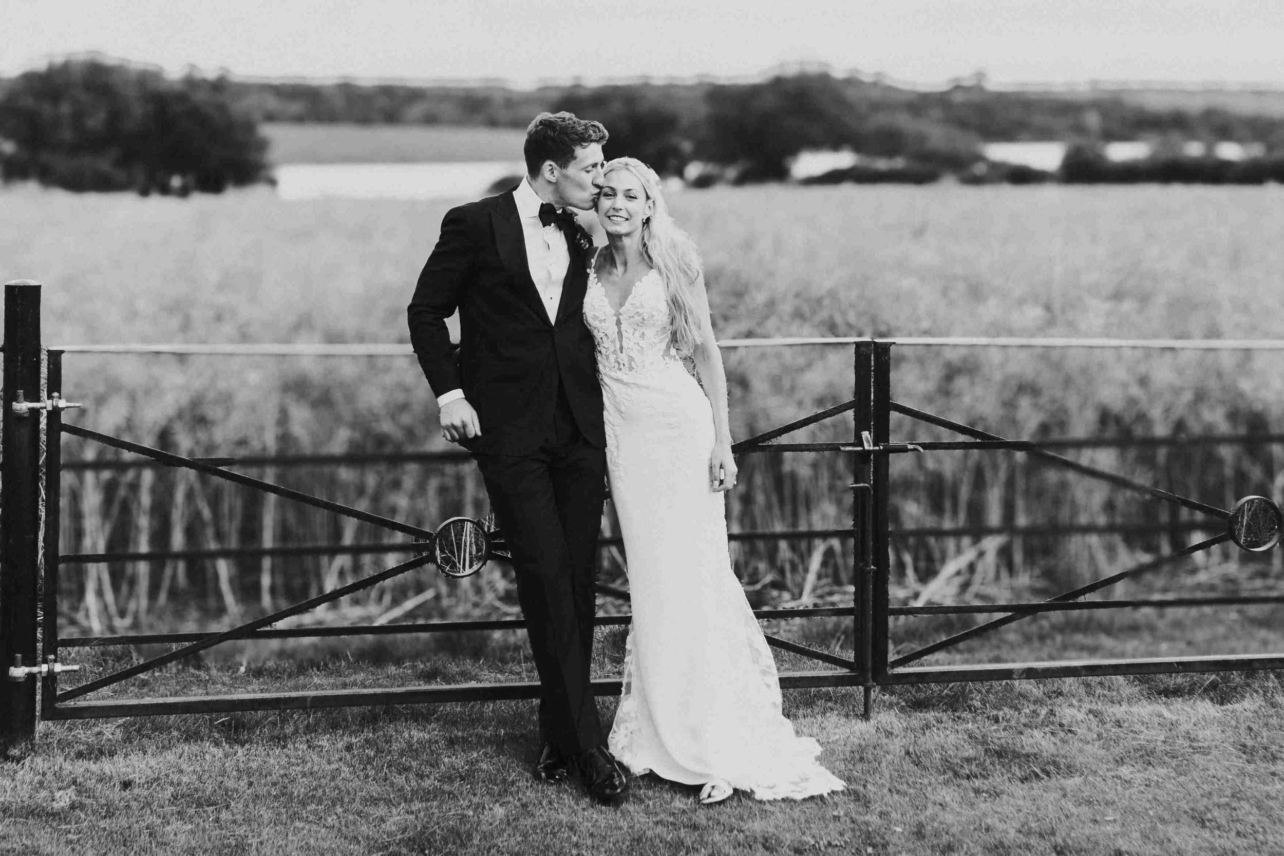 Couple standing and leaning against a metal fence while the groom kisses the bride on side of her head at the Halfmoon Farm Wedding in Rutland