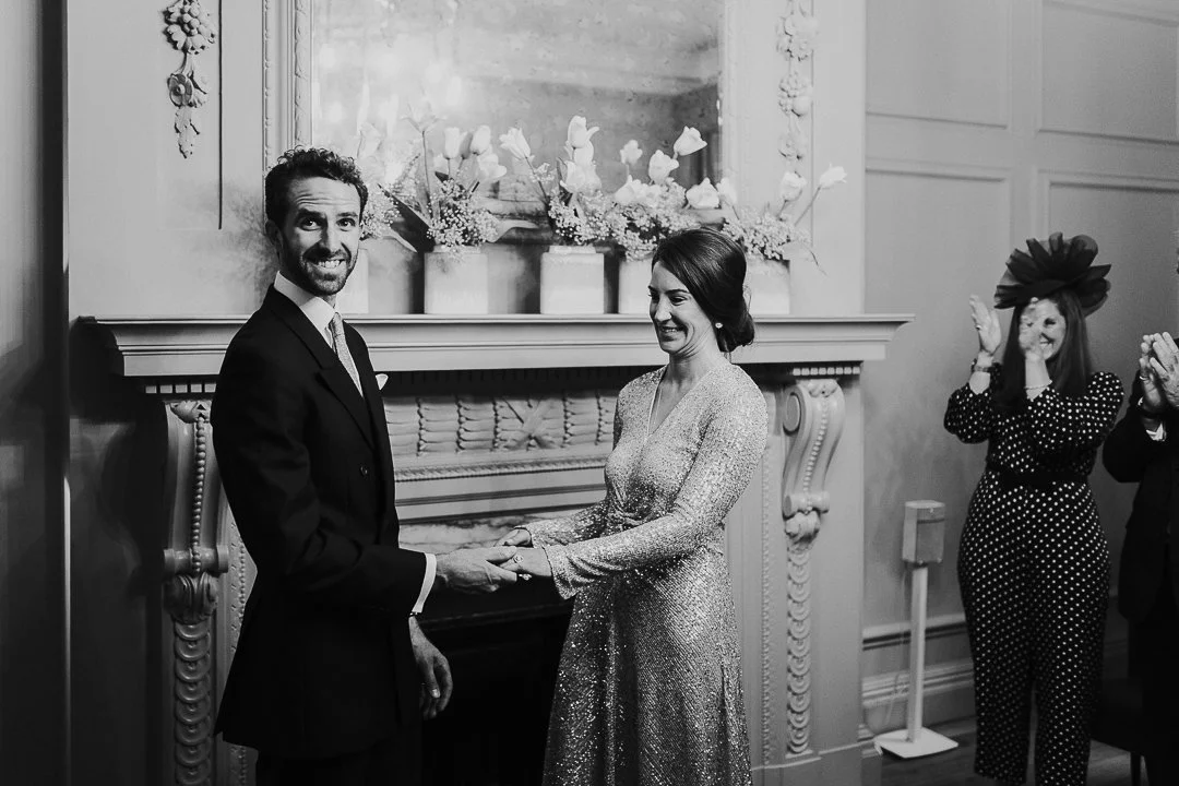 Couple holding hands while the groom looks over smiling at the camera and the bride is smiling at the groom in the ceremony at the Old Marylebone Town Hall.