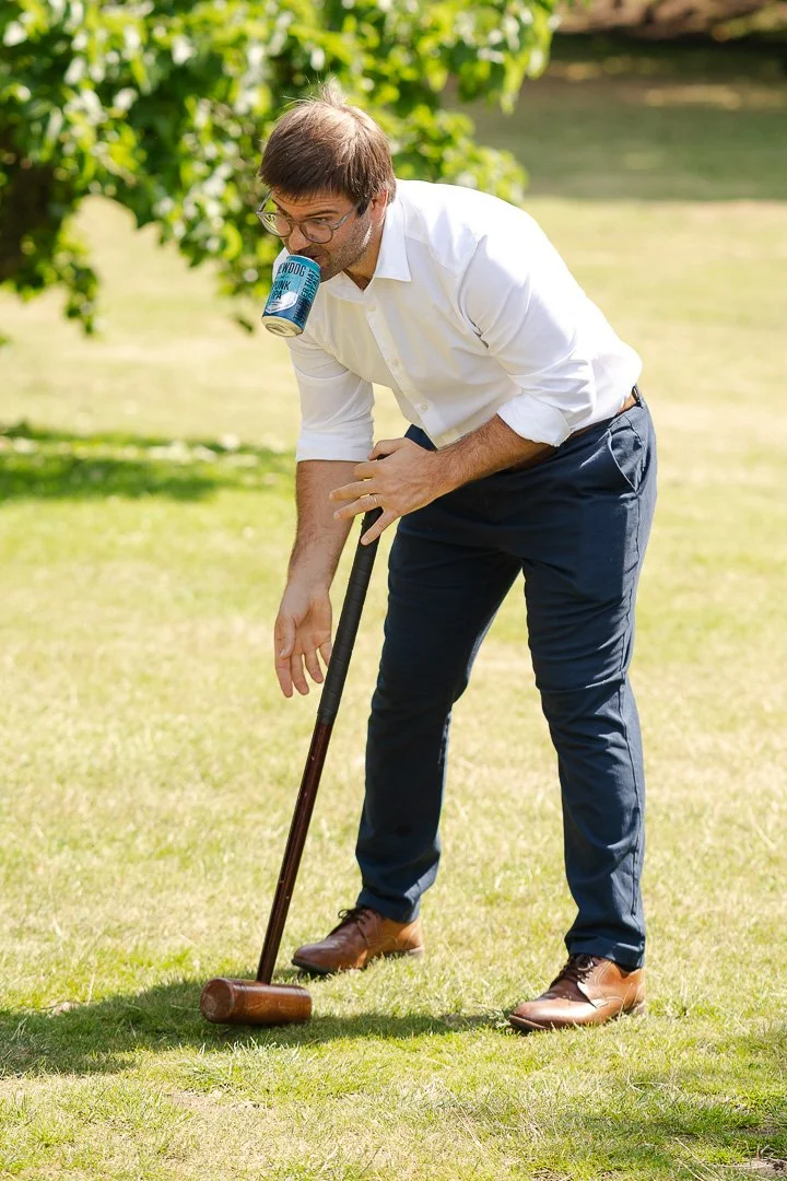 A guest holding his lager can in his mouth whilst holding his mallet playing croquet at a Hampton Court House Wedding.