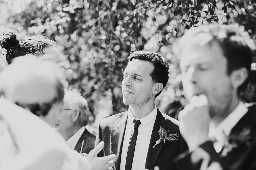 One groomsmen standing and smiling in the background whilst a guest blurred in the foreground is eating at a Hampton Court House Wedding.