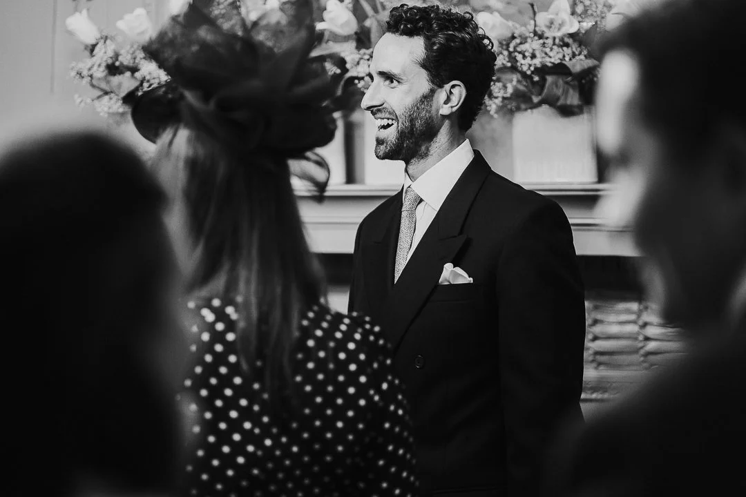 Groom standing next to fireplace smiling while bride enters the room out of shot in the ceremony room at the Old Marylebone Town Hall Wedding.