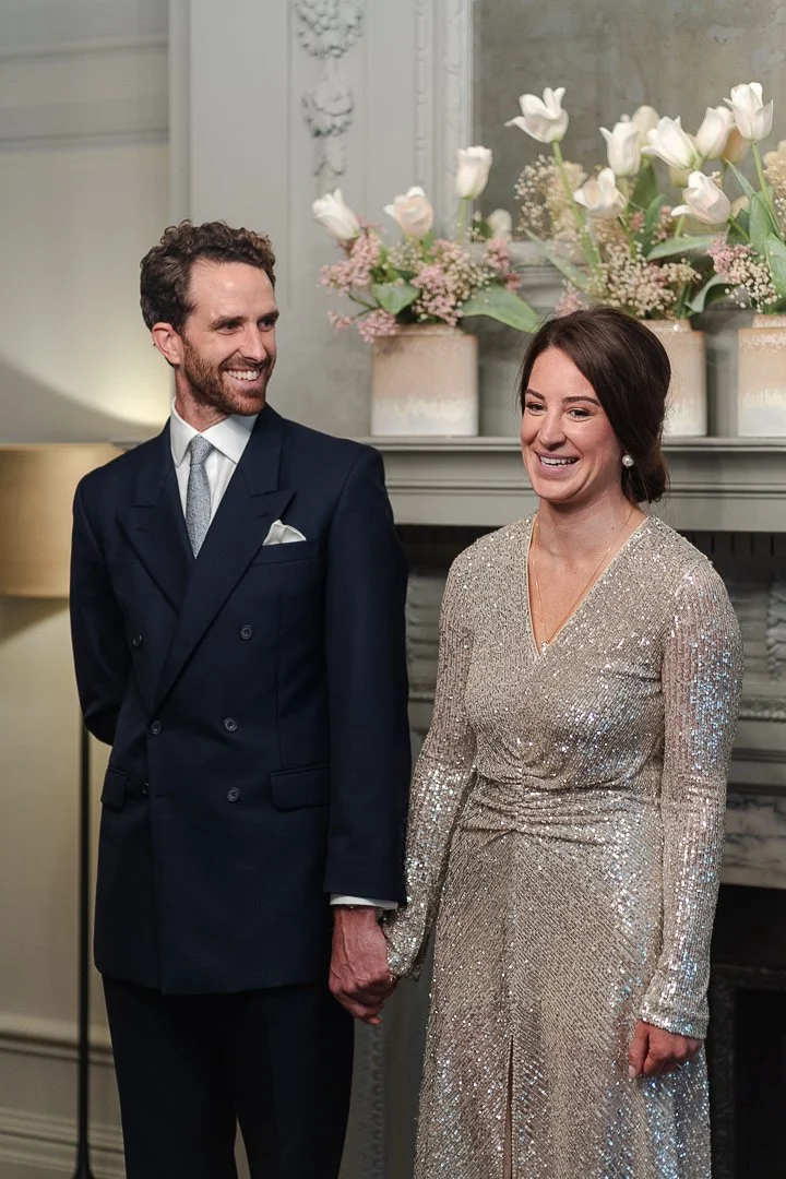 Couple holding hands while the groom smiles at the Bride as she smile looking away from him in the ceremony room at the Old Marylebone Town Hall Wedding.
